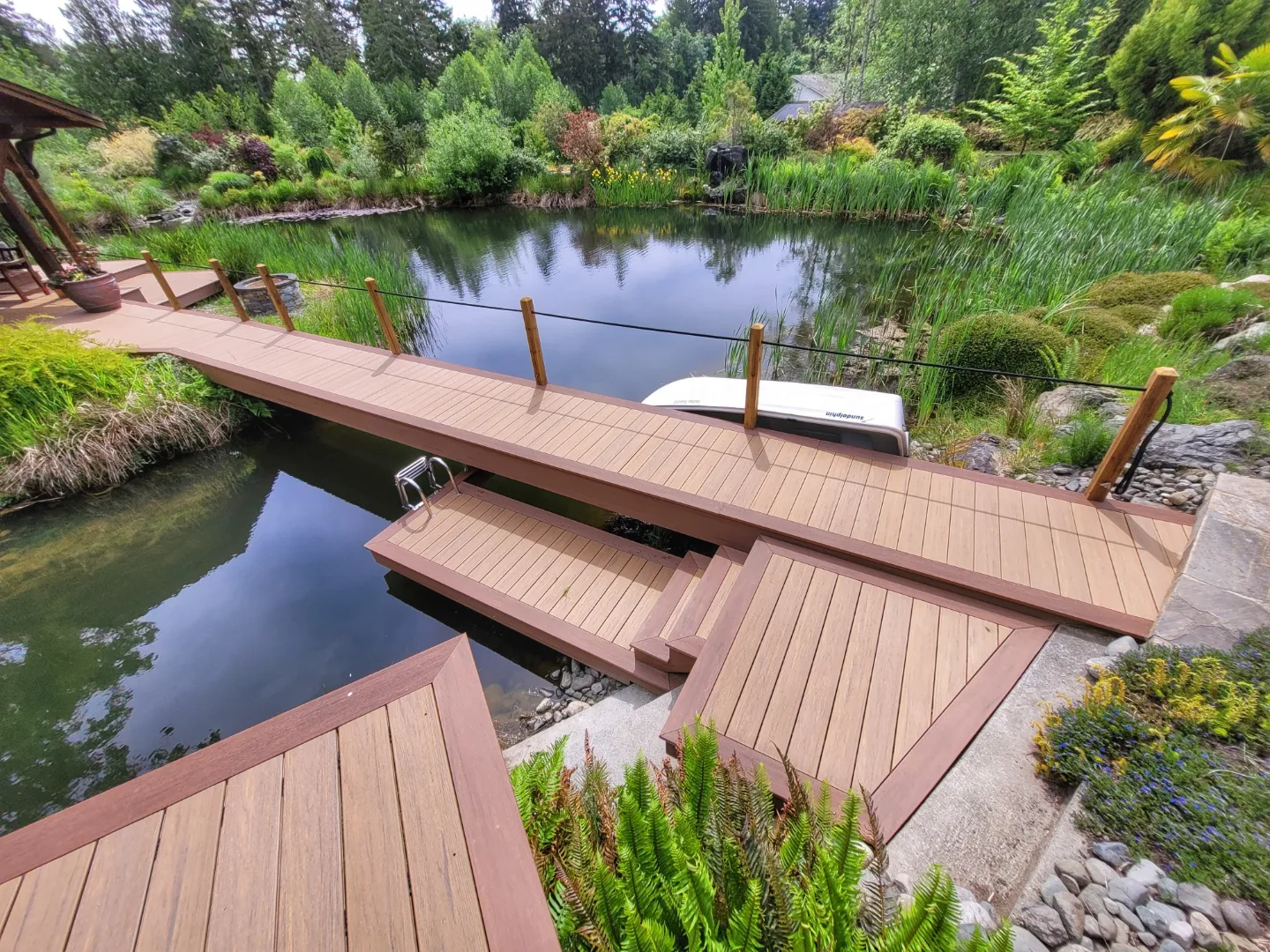Modern wooden dock with steps and railing extending over a serene garden pond surrounded by lush greenery and landscaped vegetation